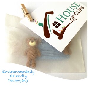 May include: A small brown teddy bear toy is packaged in a white tissue paper envelope. The envelope has a sticker with the text "The House of Clips" and a house-shaped logo. The text "Environmentally Friendly Packaging!" is written in blue on the bottom of the image.