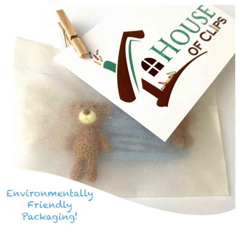 May include: A small brown teddy bear toy is packaged in a white tissue paper envelope. The envelope has a sticker with the text "The House of Clips" and a house-shaped logo. The text "Environmentally Friendly Packaging!" is written in blue on the bottom of the image.