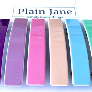 May include: Six colorful plastic hair clips in a rainbow color scheme. The clips are arranged in a row from left to right: purple, lavender, pink, peach, light blue, and mint green.