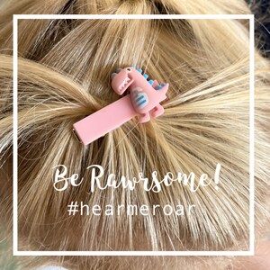 May include: A pink hair clip featuring a cartoon dinosaur with blue accents. The clip is attached to blonde hair. Text on the image reads "Be Rawrsome!" and "#hearmeroar".