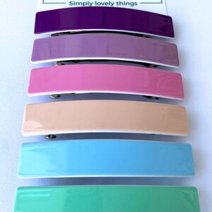 May include: Six colorful plastic hair clips in a variety of pastel colors, including purple, lavender, pink, peach, light blue, and mint green. The clips are arranged vertically, with the purple clip at the top and the mint green clip at the bottom. The clips are all the same size and shape, and they have a glossy finish.
