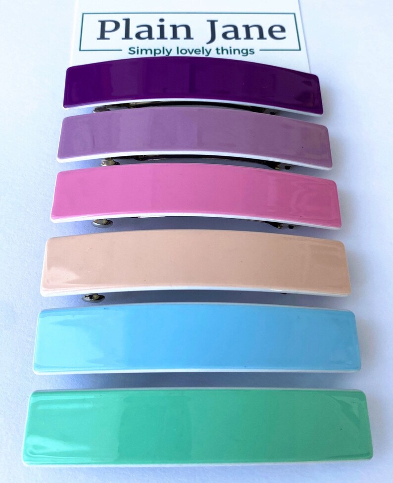 May include: Six colorful plastic hair clips in a variety of pastel colors, including purple, lavender, pink, peach, light blue, and mint green. The clips are arranged vertically, with the purple clip at the top and the mint green clip at the bottom. The clips are all the same size and shape, and they have a glossy finish.