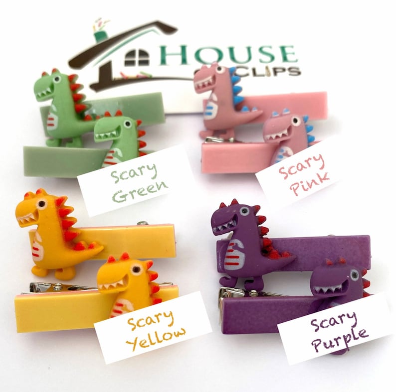 May include: Four dinosaur-themed hair clips in green, pink, yellow, and purple. Each clip features a dinosaur figure with red accents and a matching base. The clips are labeled "Scary Green", "Scary Pink", "Scary Yellow", and "Scary Purple".