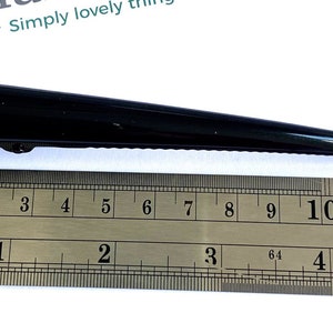 May include: A black hair clip, approximately 5 inches long, is displayed next to a ruler. The clip has a sleek, pointed design and a hinged clasp. The text "Simply lovely things" is visible in the background.