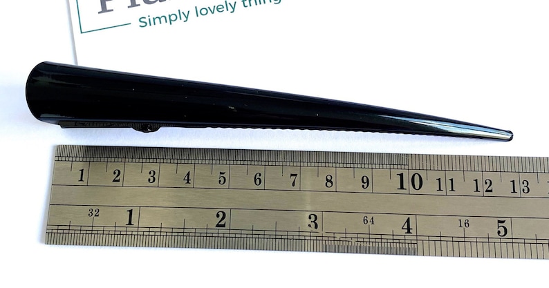 May include: A black hair clip, approximately 5 inches long, is displayed next to a ruler. The clip has a sleek, pointed design and a hinged clasp. The text "Simply lovely things" is visible in the background.