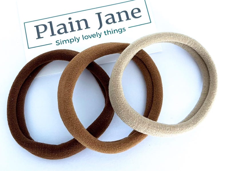 May include: Three hair scrunchies in brown, dark brown, and beige. The scrunchies are made of soft, stretchy fabric.