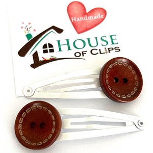以下が含まれることがあります： 縁に白いステッチのデザインが施された、茶色の木製ボタンのヘアクリップが2つ。クリップは銀色で、3つの歯があります。