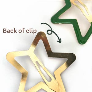 Pu&ograve; includere: Fermagli per capelli a forma di stella, dorati e verdi. Uno dei fermagli ha la forma di una stella dorata con un rivestimento in smalto verde. L'immagine mostra il retro di un fermaglio, con una freccia che lo indica. Il testo "Back of clip" &egrave; visibile.