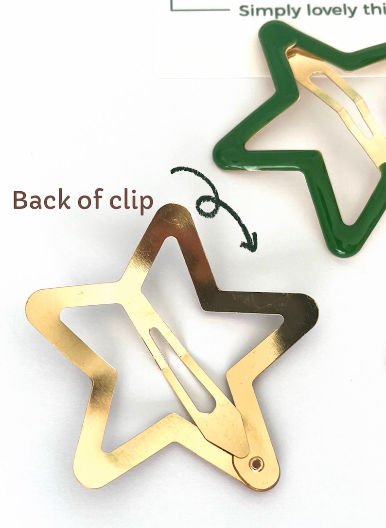 Pu&ograve; includere: Fermagli per capelli a forma di stella, dorati e verdi. Uno dei fermagli ha la forma di una stella dorata con un rivestimento in smalto verde. L'immagine mostra il retro di un fermaglio, con una freccia che lo indica. Il testo "Back of clip" &egrave; visibile.