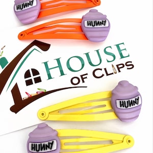 Könnte beinhalten: Vier Haarspangen mit Honigtopf-Design. Zwei Spangen sind orange mit lavendelfarbenen Honigtöpfen, und zwei sind gelb mit lavendelfarbenen Honigtöpfen. Die Honigtöpfe tragen das Wort "HUNNY" in schwarzen Buchstaben. Das Logo "HOUSE OF CLIPS" ist im Hintergrund.