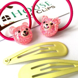 May include: Two pink hair ties with sparkly pink bear charms and two yellow plastic hair clips. The hair ties and clips are on a white surface with a white card that says "House of Clips".