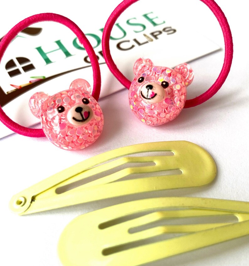 May include: Two pink hair ties with sparkly pink bear charms and two yellow plastic hair clips. The hair ties and clips are on a white surface with a white card that says "House of Clips".