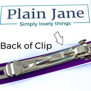 May include: Back of a purple and white plastic hair clip with a silver metal clasp. The clip is designed to hold hair in place.