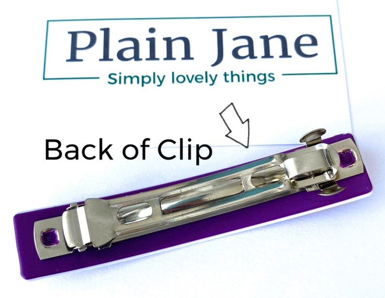 May include: Back of a purple and white plastic hair clip with a silver metal clasp. The clip is designed to hold hair in place.