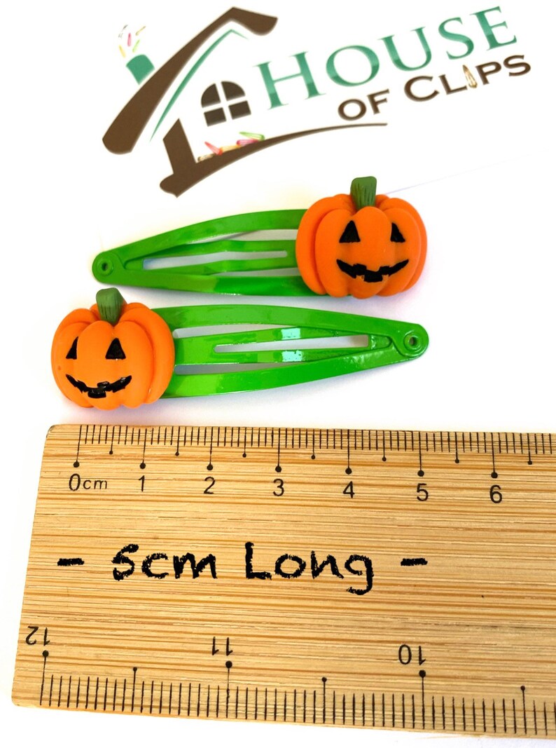 Op de afbeelding: Twee groene haarclips met oranje pompoen-vormige bedels. De pompoenen hebben zwarte ogen, een neus en een glimlachende mond. De clips zijn 5 cm lang.