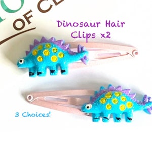 May include: Two hair clips featuring bright blue dinosaur figures with purple dorsal plates and yellow spots. The dinosaurs are attached to light pink metal snap clips. Text on the image reads Dinosaur Hair Clips x2 and 3 Choices!