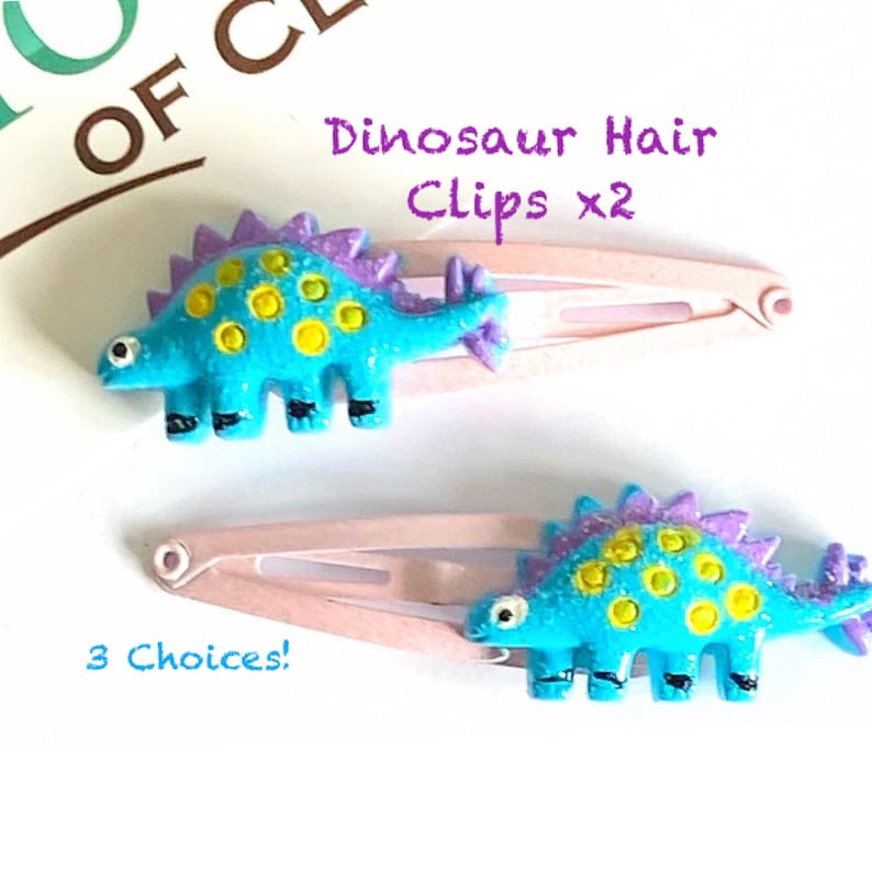 May include: Two hair clips featuring bright blue dinosaur figures with purple dorsal plates and yellow spots. The dinosaurs are attached to light pink metal snap clips. Text on the image reads Dinosaur Hair Clips x2 and 3 Choices!