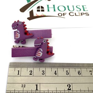 May include: Two purple dinosaur hair clips. Each clip features a cartoon dinosaur with red accents and white stripes. The clips are approximately 2 inches long, as measured by the ruler in the image. The clips are from House of Clips.