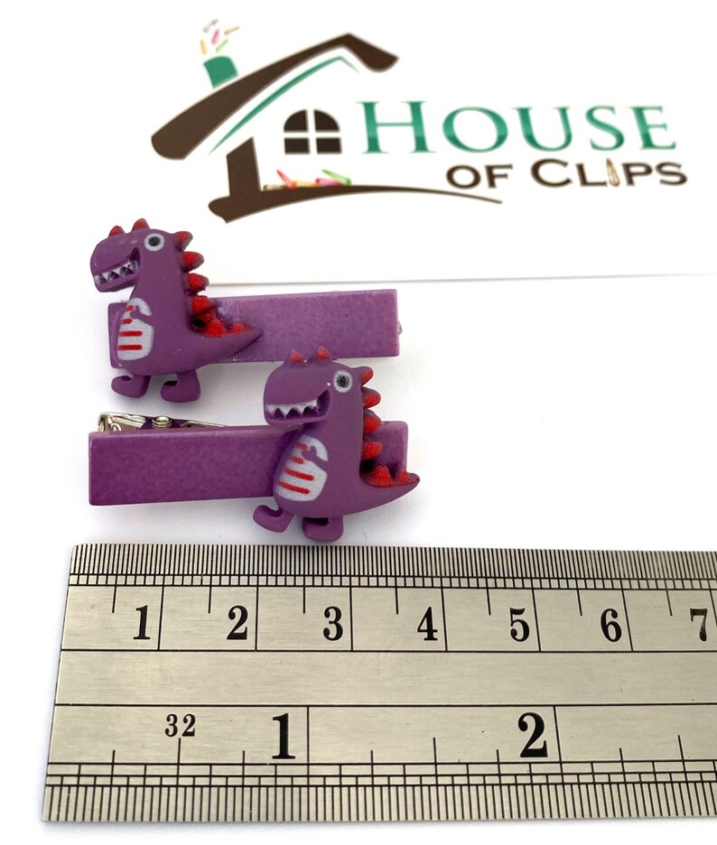 May include: Two purple dinosaur hair clips. Each clip features a cartoon dinosaur with red accents and white stripes. The clips are approximately 2 inches long, as measured by the ruler in the image. The clips are from House of Clips.
