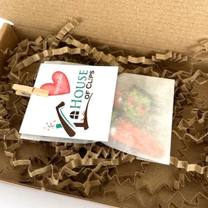 Pode incluir: Um pequeno cart&atilde;o branco com o texto "The House of Clips" e um cora&ccedil;&atilde;o vermelho com a palavra "Handmade" nele. O cart&atilde;o est&aacute; em uma caixa de papel&atilde;o marrom com material de embalagem de papel marrom.