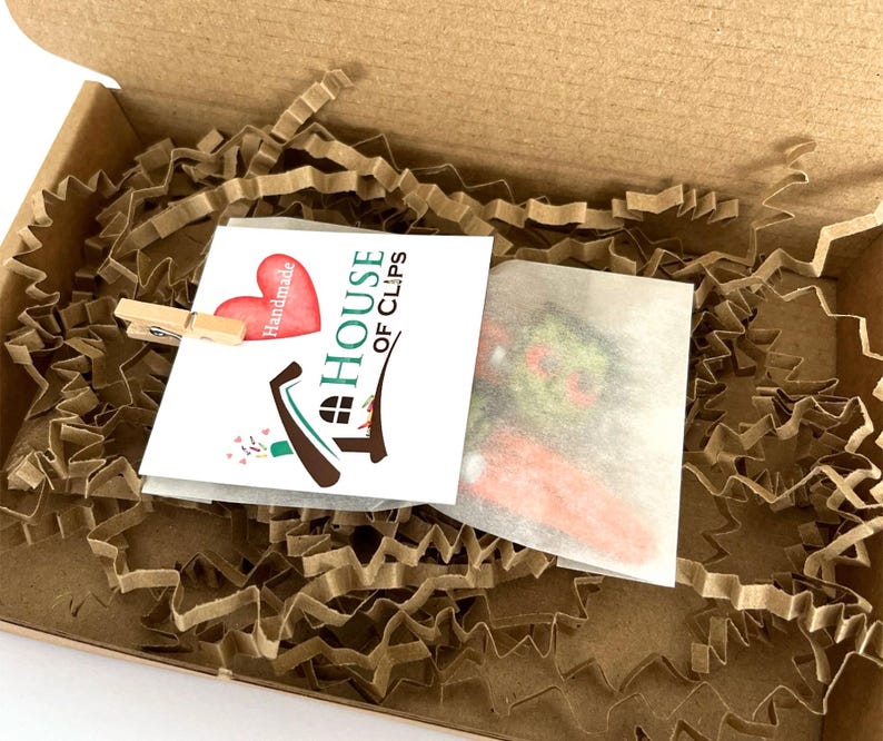 Pode incluir: Um pequeno cart&atilde;o branco com o texto "The House of Clips" e um cora&ccedil;&atilde;o vermelho com a palavra "Handmade" nele. O cart&atilde;o est&aacute; em uma caixa de papel&atilde;o marrom com material de embalagem de papel marrom.