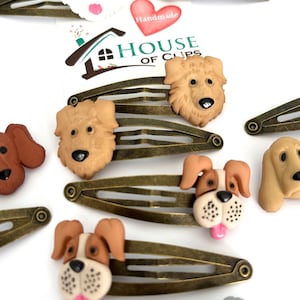 Könnte beinhalten: Eine Sammlung von Haarspangen aus bronzefarbenem Metall, die jeweils mit einem detaillierten, dreidimensionalen Hundekopf verziert sind. Die Hundeköpfe variieren in Rasse und Farbe, darunter Weiß, Braun und Hellbraun. Das Bild zeigt auch einen herzförmigen Aufkleber mit dem Wort "Handmade".