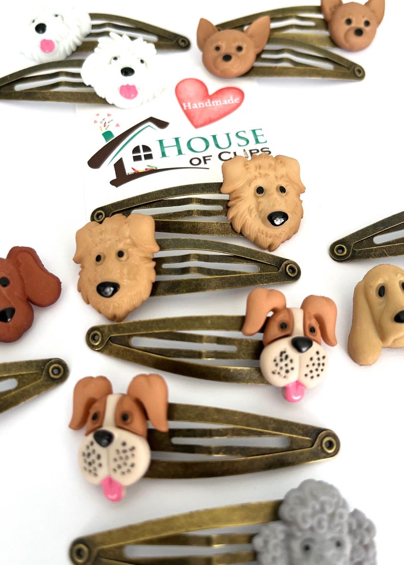 May include: A collection of bronze-toned metal hair clips, each adorned with a detailed, three-dimensional dog head. The dog heads vary in breed and color, including white, brown, and tan. The image also features a heart-shaped sticker with the word "Handmade".