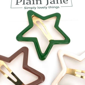 Pu&ograve; includere: Fermagli per capelli a forma di stella nei colori verde, marrone e crema. Ogni fermaglio ha una chiusura in metallo color oro. Il marchio "Plain Jane" &egrave; stampato nella parte superiore dell'immagine.
