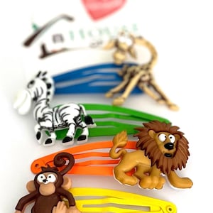 Könnte beinhalten: Eine Sammlung bunter Haarspangen mit Tiermotiven. Die Spangen zeigen ein Zebra, eine Giraffe, einen Löwen und einen Affen, jeweils an einer andersfarbigen Spange: blau, grün, orange und gelb. Die Spangen sind für Kinder konzipiert.