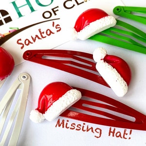 May include: Four red and white Santa hat hair clips, two on red clips, one on a green clip, and one on a silver clip. The text "Santa's" is visible on a white background. The text "Missing Hat!" is written on a red clip.