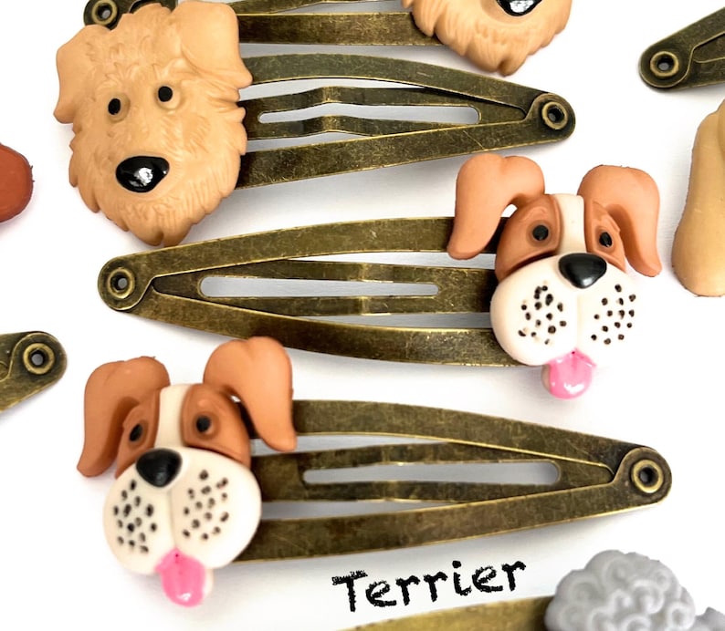 May include: Several bronze-toned metal hair clips with dog-shaped embellishments. The dog faces are tan, brown, and white, with black eyes and pink tongues. The word "Terrier" is printed in black.