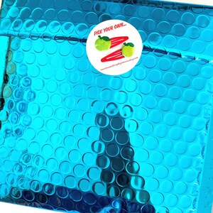K&ouml;nnte beinhalten: Ein blauer Luftpolsterumschlag mit einem wei&szlig;en runden Aufkleber, auf dem "Pick your own..." steht und ein Bild von zwei gr&uuml;nen Apfel-Haarklammern mit roten Haarklammern zu sehen ist. Die Website-Adresse "www.houseofklippityklop.etsy.com" ist auf dem Aufkleber aufgedruckt.