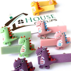 May include: A collection of colorful dinosaur hair clips. The clips feature small, cartoon-style dinosaurs in shades of green, pink, yellow, and purple, with red and blue accents. Each dinosaur is attached to a rectangular barrette clip.