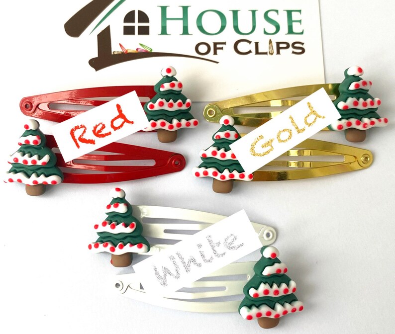 K&ouml;nnte beinhalten: Drei Haarklammern mit Weihnachtsbaum-Anh&auml;ngern. Die Klammern sind rot, gold und wei&szlig;. Die Anh&auml;nger sind gr&uuml;n mit wei&szlig;en und roten Details. Der Text "Rot", "Gold" und "Wei&szlig;" steht auf wei&szlig;en Etiketten.