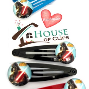 May include: A selection of hair clips in blue, black, and red, each featuring a Star Wars design with Darth Vader and a lightsaber. The clips have a round image and are set against a white backdrop. The image also includes a heart with the word "Handmade".