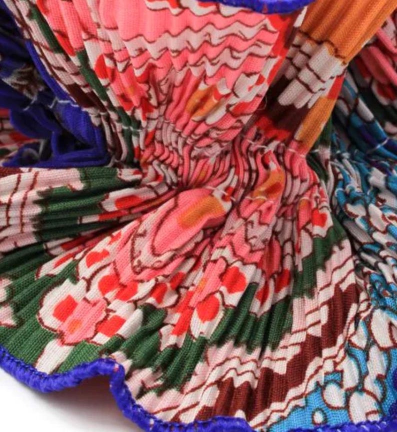 May include: Close-up of a pleated fabric with a colorful floral pattern. The fabric is pink, orange, green, blue, and white. The fabric has a blue trim.