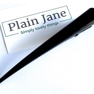 May include: A black, pointed hair clip with a wide, rounded top and a hinged clip. The clip is positioned next to a white card with the text "Plain Jane" and "Simply lovely things" in a green border.