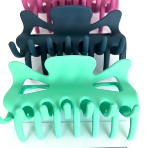 May include: A stack of colorful hair claw clips in shades of pink, purple, teal, and navy blue. The clips are arranged on a white surface, with the brand name "Plain Jane" and the tagline "Simply lovely things" visible at the bottom.