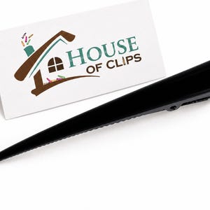 May include: A long, sleek black hair clip with a textured edge, positioned next to a white rectangular card featuring the brown and teal logo for House of Clips, which depicts a stylized house with a chimney and small colorful clips.