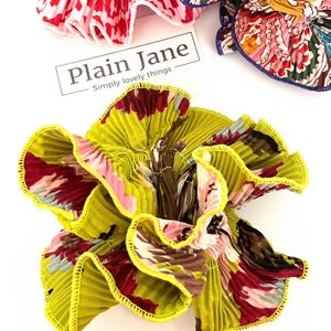 May include: A yellow, green, pink, and brown floral fabric hair scrunchie with a yellow ruffled edge. The scrunchie is made of pleated fabric and has a  "Plain Jane Simply lovely things" label.