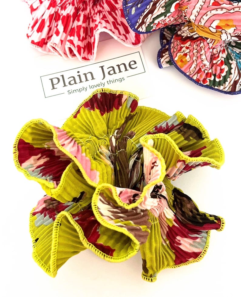 May include: A yellow, green, pink, and brown floral fabric hair scrunchie with a yellow ruffled edge. The scrunchie is made of pleated fabric and has a  "Plain Jane Simply lovely things" label.