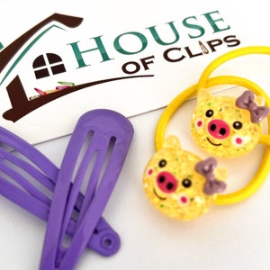 May include: Two yellow pig hair ties with purple bows and two purple hair clips. The hair ties and clips are on a white surface with a card that says "HOUSE OF CLIPS".