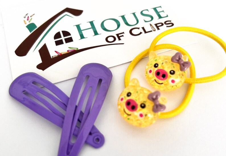 May include: Two yellow pig hair ties with purple bows and two purple hair clips. The hair ties and clips are on a white surface with a card that says "HOUSE OF CLIPS".