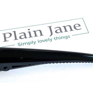 May include: A glossy black hair clip with a pointed end and a hinged clip. The clip has a ridged edge and is set against a white background. The text "Plain Jane Simply lovely things" is visible.