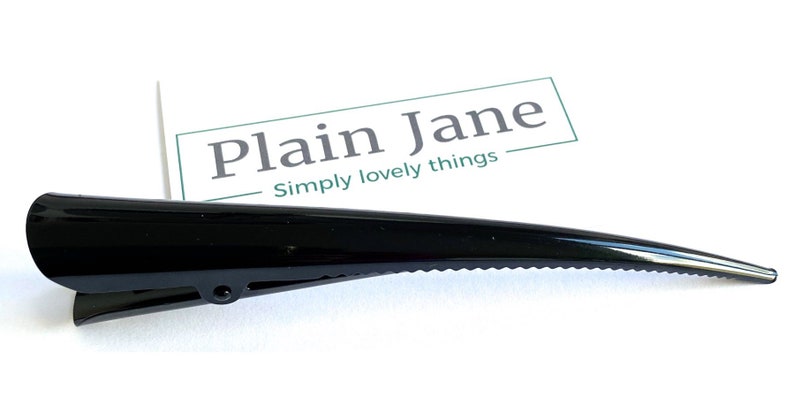 May include: A glossy black hair clip with a pointed end and a hinged clip. The clip has a ridged edge and is set against a white background. The text "Plain Jane Simply lovely things" is visible.