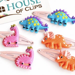 May include: Five hair clips featuring miniature dinosaur figures in pink, blue, and orange with glittery finishes. The clips are attached to a card labeled House of Clips. The dinosaurs include long-necked sauropods, spiky-backed stegosaurs, and three-horned triceratops designs.