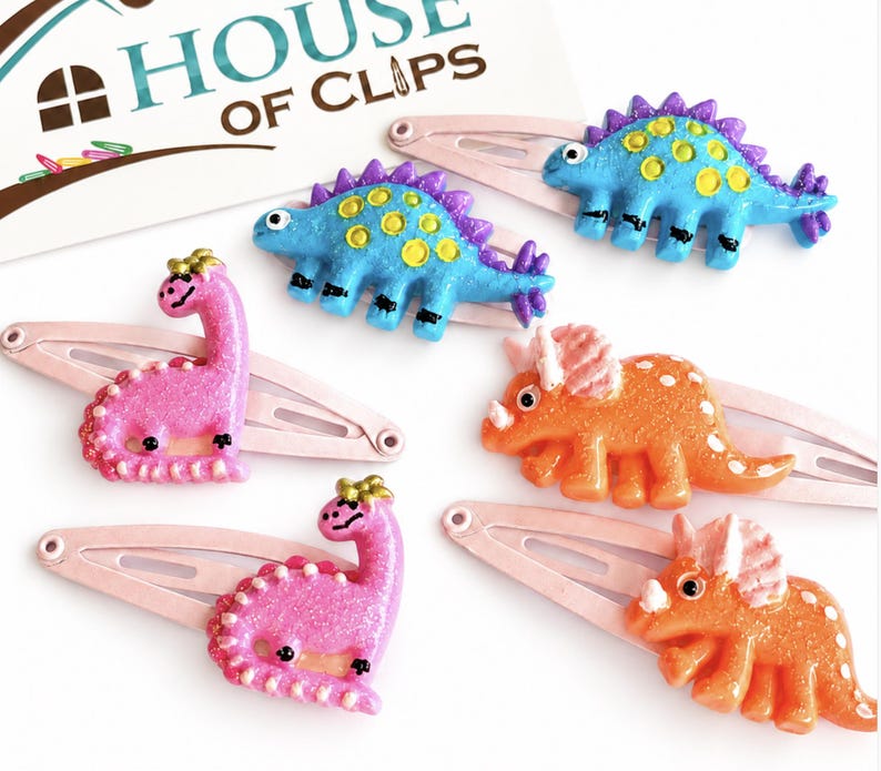 May include: Five hair clips featuring miniature dinosaur figures in pink, blue, and orange with glittery finishes. The clips are attached to a card labeled House of Clips. The dinosaurs include long-necked sauropods, spiky-backed stegosaurs, and three-horned triceratops designs.