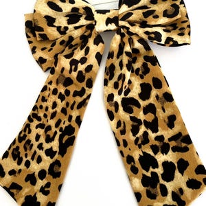May include: A large leopard print hair bow. The bow features a tan background with black spots and long tails. The text "Plain Jane - Simply lovely things" is displayed on a white card.