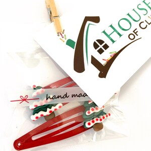 K&ouml;nnte beinhalten: Zwei rote Haarklammern mit gr&uuml;nen und wei&szlig;en Christbaumschmuck. Die Ornamente haben rote und wei&szlig;e Zuckerstangenakzente. Die Verpackung enth&auml;lt ein kleines wei&szlig;es Etikett mit dem Text "hand made" in Schwarz.