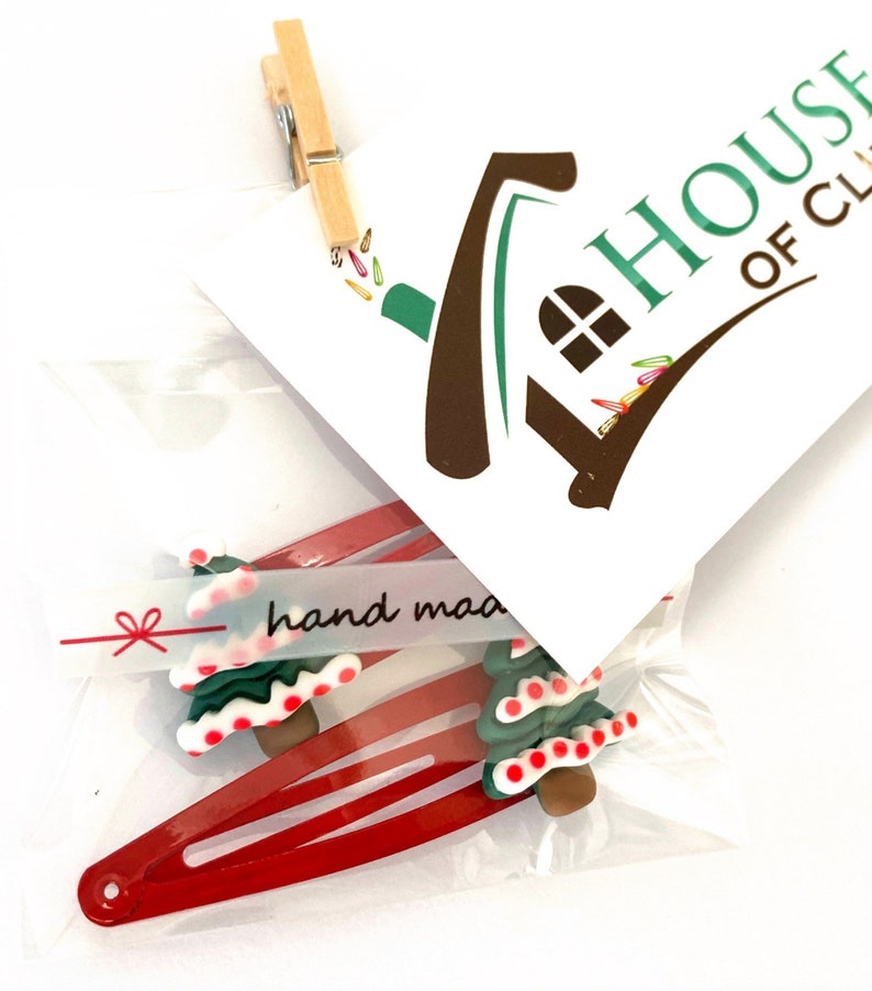 K&ouml;nnte beinhalten: Zwei rote Haarklammern mit gr&uuml;nen und wei&szlig;en Christbaumschmuck. Die Ornamente haben rote und wei&szlig;e Zuckerstangenakzente. Die Verpackung enth&auml;lt ein kleines wei&szlig;es Etikett mit dem Text "hand made" in Schwarz.